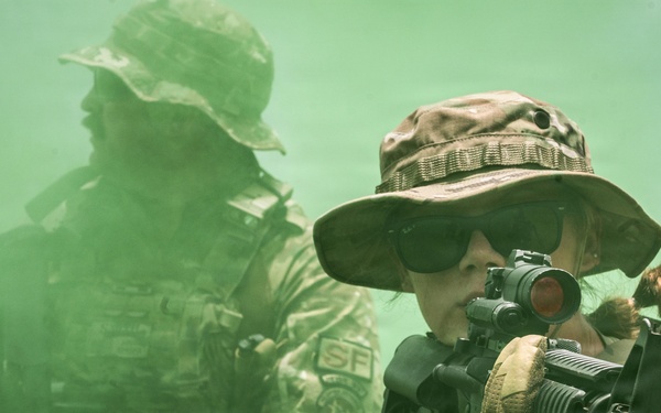 Two armed individuals in tactical military-style gear are moving through dense green smoke. The foreground figure, wearing a boonie hat and sunglasses, is aiming a rifle equipped with a red dot optic. Both individuals wear camouflage uniforms and carry tactical equipment including backpacks and weapons.