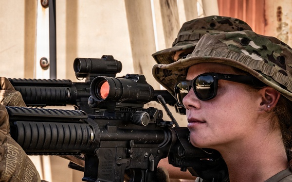 A female military or law enforcement personnel is shown in a tactical shooting stance, aiming an M4/AR-15 style rifle equipped with optical sights and accessories. She is wearing multicam camouflage gear including a boonie hat, tactical gloves branded 'Mechanix', sunglasses, and a plate carrier vest. The image appears to be taken during a training exercise or field operation.