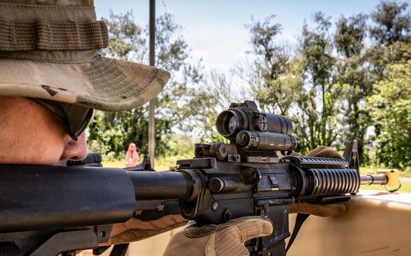 A military or law enforcement personnel is aiming an assault rifle from behind a barricade in an outdoor training or operational environment. The shooter is wearing camouflage gear, a boonie hat, tactical gloves, and eye protection. The setting appears to be a shooting range or field exercise with green vegetation visible in the background.