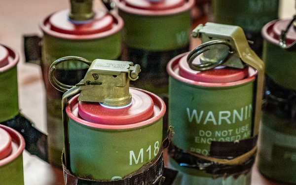The image shows multiple M18 colored smoke grenades arranged on a wooden surface. These are military/law enforcement smoke grenades with red tops and olive drab bodies, several secured with black tape. Visible markings include 'M18', 'RED', lot numbers such as 'PB-11M405', and warning text reading 'WARNING DO NOT USE ENCLOSED A...'