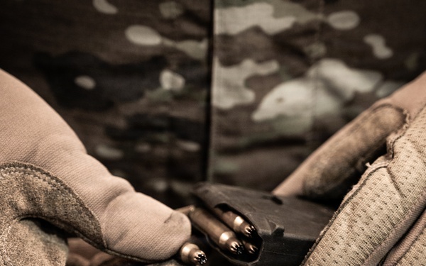 A person wearing tactical/military gloves and camouflage clothing is loading ammunition rounds into a firearm magazine. The close-up shot focuses on the hands and the loading action, with a shallow depth of field creating a blurred background. The image has a desaturated, sepia-toned aesthetic typical of military or tactical photography.