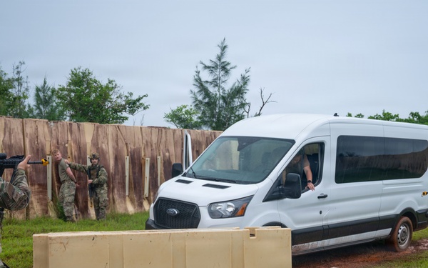 The image depicts a military training exercise at what appears to be a simulated checkpoint or entry control point. Armed personnel in multicam/OCP camouflage uniforms are conducting a vehicle stop procedure on a white Ford Transit van. Concrete barriers and a wooden wall structure are present, consistent with a purpose-built training facility.