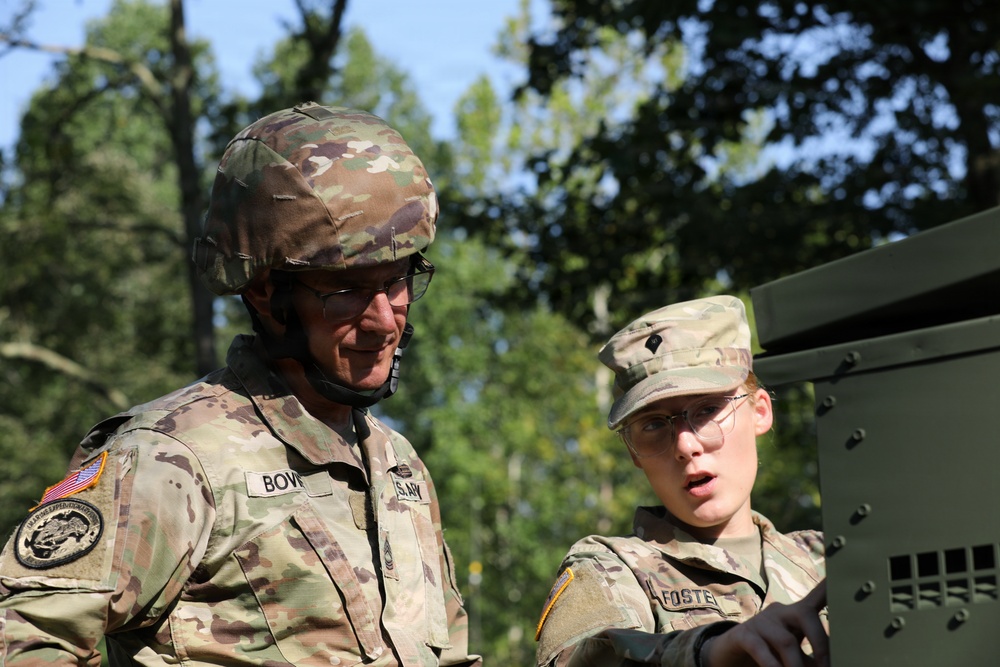 Two U.S. Army soldiers in OCP (Operational Camouflage Pattern) uniforms are engaged in what appears to be a training or operational activity near a military vehicle. The soldier on the left, identified by the name tape 'BOVE', is wearing a combat helmet and glasses, while the soldier on the right, identified as 'FOSTER', is wearing a patrol cap and safety glasses. Both soldiers appear to be examining or operating equipment on a green military vehicle.