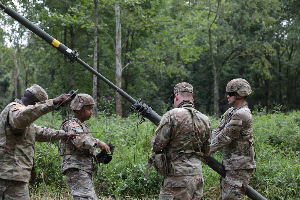 Four U.S. Army soldiers in Operational Camouflage Pattern (OCP) uniforms are working together to erect or handle a tall antenna mast in a wooded, vegetated area. The soldiers appear to be conducting a field communications setup exercise or training operation. At least one soldier's name tape ('BOCKOV' or similar) is partially visible on their uniform.