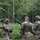 Four U.S. Army soldiers in Operational Camouflage Pattern (OCP) uniforms are working together to erect or handle a tall antenna mast in a wooded, vegetated area. The soldiers appear to be conducting a field communications setup exercise or training operation. At least one soldier's name tape ('BOCKOV' or similar) is partially visible on their uniform.