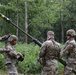 Four U.S. Army soldiers in Operational Camouflage Pattern (OCP) uniforms are working together to erect or handle a tall antenna mast in a wooded, vegetated area. The soldiers appear to be conducting a field communications setup exercise or training operation. At least one soldier's name tape ('BOCKOV' or similar) is partially visible on their uniform.