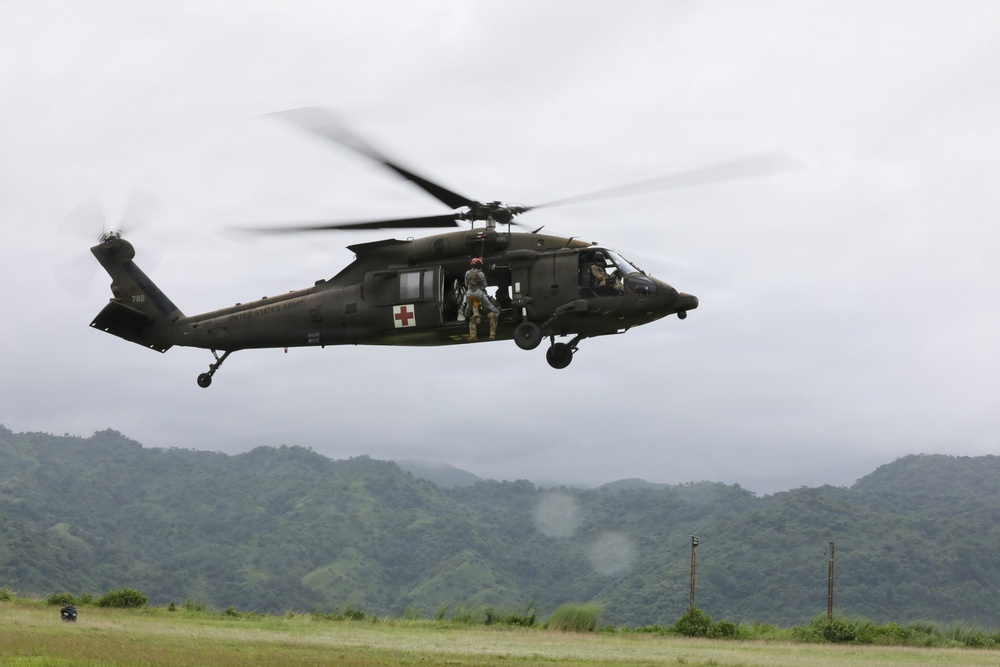 A U.S. Army UH-60 Black Hawk helicopter configured as a medevac (medical evacuation) aircraft is hovering at low altitude over a grassy field. The helicopter displays a red cross emblem indicating its medical role and bears 'United States Army' markings with tail number 782. The backdrop features lush, green mountainous terrain under an overcast sky, suggesting a tropical or subtropical environment.