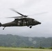A U.S. Army UH-60 Black Hawk helicopter configured as a medevac (medical evacuation) aircraft is hovering at low altitude over a grassy field. The helicopter displays a red cross emblem indicating its medical role and bears 'United States Army' markings with tail number 782. The backdrop features lush, green mountainous terrain under an overcast sky, suggesting a tropical or subtropical environment.