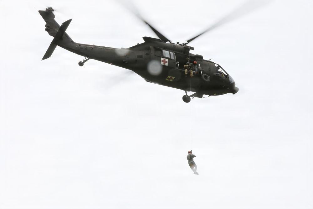 A dark olive/black military medical helicopter is airborne against an overcast sky, with a person suspended below it on a hoist or cable. The helicopter displays a red cross emblem and a white cross insignia, identifying it as a MEDEVAC (medical evacuation) aircraft. This appears to be a hoist rescue or training exercise in progress.