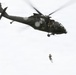 A dark olive/black military medical helicopter is airborne against an overcast sky, with a person suspended below it on a hoist or cable. The helicopter displays a red cross emblem and a white cross insignia, identifying it as a MEDEVAC (medical evacuation) aircraft. This appears to be a hoist rescue or training exercise in progress.
