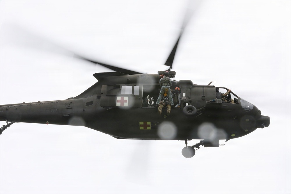 A United States Army medical evacuation (MEDEVAC) helicopter, identifiable as a UH-60 Black Hawk, is captured in flight with its side door open. Personnel are visible hanging or leaning out of the open cabin door, suggesting a rescue or hoist operation in progress. Red cross markings on the fuselage confirm its medical/MEDEVAC designation.