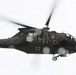 A United States Army medical evacuation (MEDEVAC) helicopter, identifiable as a UH-60 Black Hawk, is captured in flight with its side door open. Personnel are visible hanging or leaning out of the open cabin door, suggesting a rescue or hoist operation in progress. Red cross markings on the fuselage confirm its medical/MEDEVAC designation.