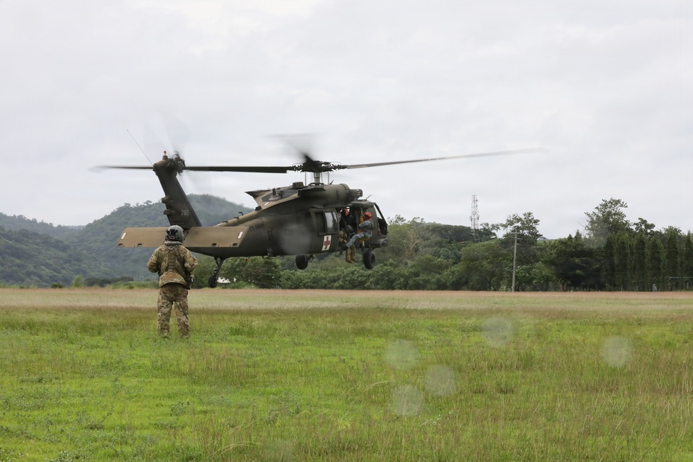 A U.S. Army UH-60 Black Hawk helicopter marked with a Red Cross emblem is hovering low over a grassy field, indicating a medical evacuation (MEDEVAC) operation. A soldier in full combat gear stands in the foreground observing the aircraft, while personnel are visible in the open cabin door. The setting appears to be a tropical or subtropical region based on the lush green vegetation and mountainous background.
