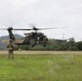 A U.S. Army UH-60 Black Hawk helicopter marked with a Red Cross emblem is hovering low over a grassy field, indicating a medical evacuation (MEDEVAC) operation. A soldier in full combat gear stands in the foreground observing the aircraft, while personnel are visible in the open cabin door. The setting appears to be a tropical or subtropical region based on the lush green vegetation and mountainous background.