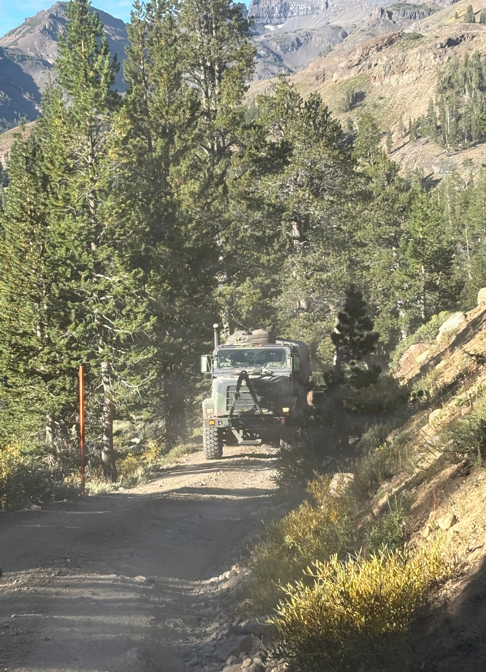 A large military-style cargo truck is traveling down a narrow, unpaved mountain dirt road surrounded by conifer trees. The rugged alpine terrain features rocky slopes, sparse high-altitude vegetation, and dramatic mountain peaks visible in the background. The scene appears to be at high elevation, likely in a western U.S. mountain range such as the Sierra Nevada or Rocky Mountains.