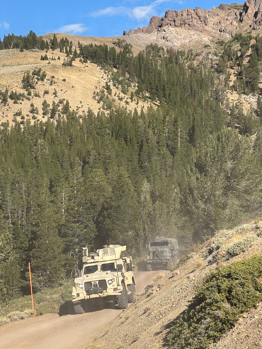 Two military vehicles are traveling along a narrow dirt mountain road in a rugged, forested alpine environment. The lead vehicle appears to be a tan/desert-colored Joint Light Tactical Vehicle (JLTV) or similar MRAP-type vehicle, followed by a larger olive-drab military truck. The convoy is kicking up dust as it navigates the steep terrain surrounded by conifer trees and rocky mountain peaks.