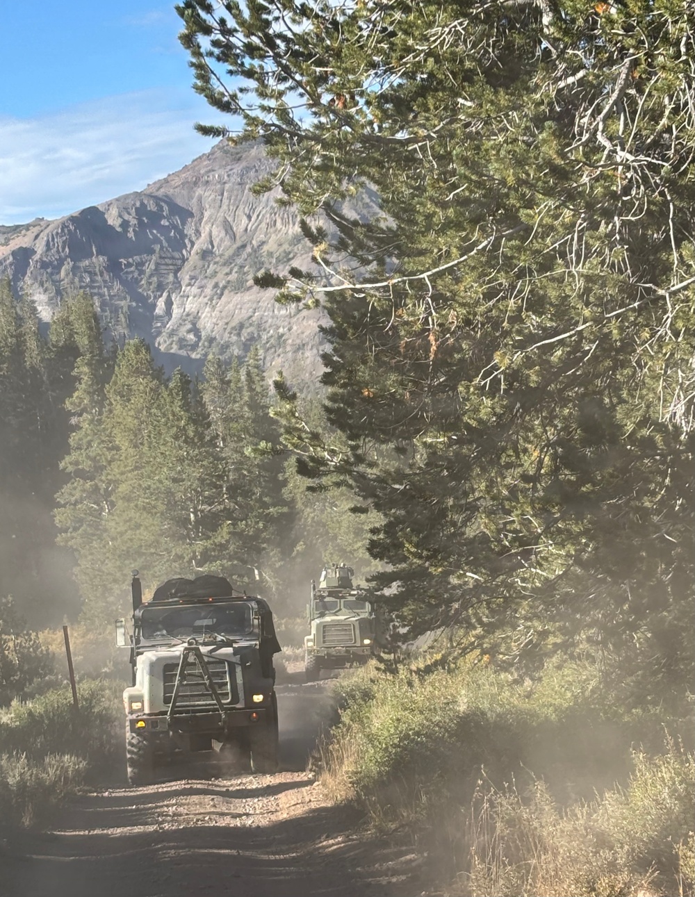 Two military-style heavy trucks are traveling in convoy along a narrow, unpaved dirt road through a mountainous forested area. The vehicles are kicking up significant dust as they navigate the trail lined with conifer trees. A large rocky mountain peak is visible in the background under a partly cloudy blue sky.