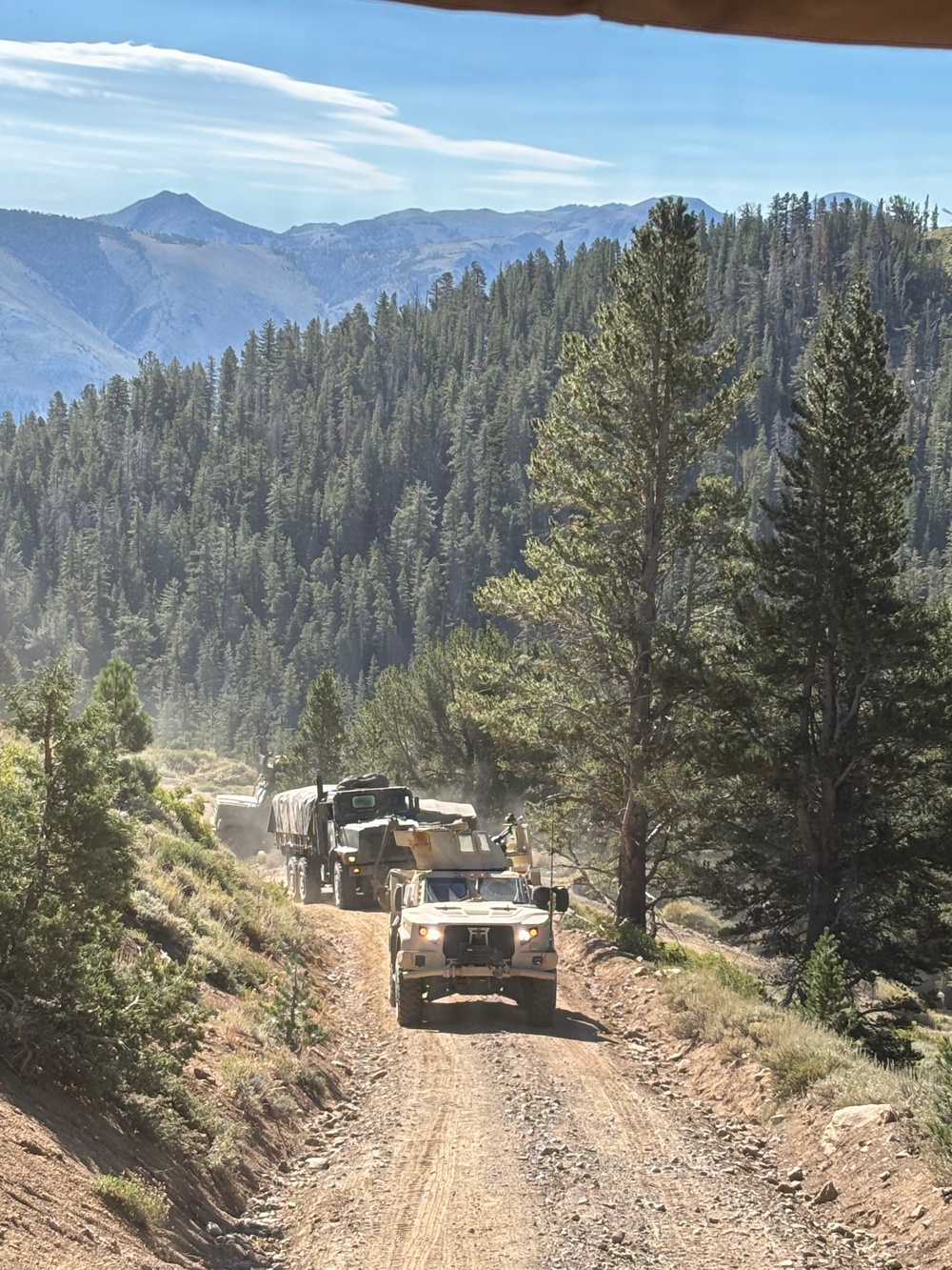 A military convoy of tactical vehicles is traveling along a narrow dirt mountain road surrounded by dense conifer forest. The lead vehicle appears to be an JLTV (Joint Light Tactical Vehicle) or similar MRAP-type vehicle, followed by at least two larger military cargo trucks. The setting appears to be a high-altitude mountainous region, likely in the western United States based on the terrain and vegetation.