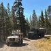 Two military vehicles are traveling along a dirt/gravel road through a coniferous forest under clear blue skies. The convoy consists of a tan/desert-colored Joint Light Tactical Vehicle (JLTV) in the lead and an olive drab Medium Tactical Vehicle (MTV/LMTV) cargo truck following behind. At least one soldier is visible manning a turret position on the lead vehicle.