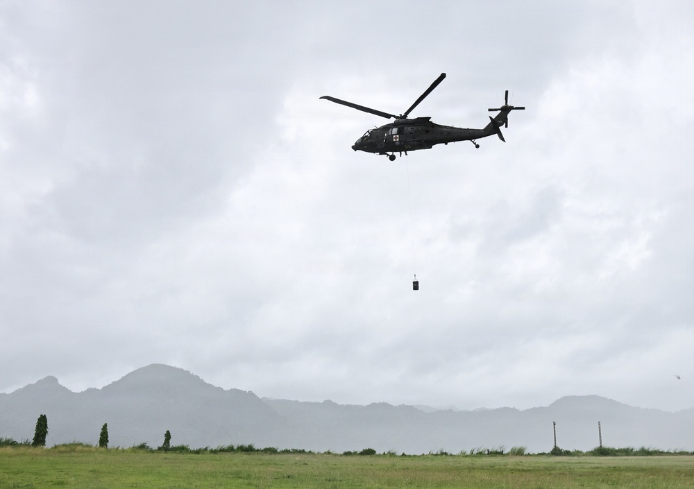 A military helicopter, identifiable by a cross/medical marking on its fuselage, is hovering over a grassy field while suspending a dark-colored load on a cable beneath it. The scene appears to be a sling-load or hoist operation conducted in overcast, possibly rainy conditions. A mountainous landscape is visible in the misty background.