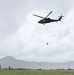 A military helicopter, identifiable by a cross/medical marking on its fuselage, is hovering over a grassy field while suspending a dark-colored load on a cable beneath it. The scene appears to be a sling-load or hoist operation conducted in overcast, possibly rainy conditions. A mountainous landscape is visible in the misty background.