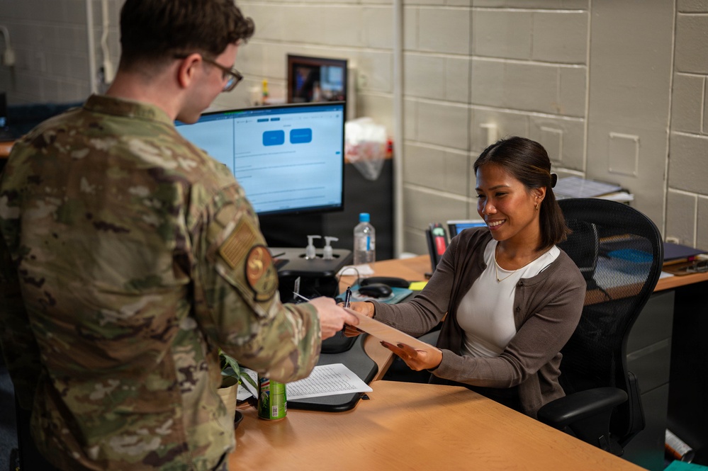 A military service member in camouflage uniform is handing a document to a civilian woman seated at an office desk. The woman is smiling warmly as she receives the paperwork. The setting appears to be a military administrative office.