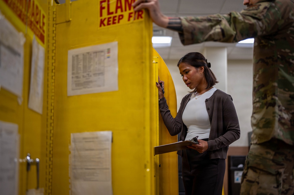 A civilian woman in business casual attire holds a clipboard and inspects or reviews a large yellow flammable materials storage cabinet. A person in military camouflage uniform (OCP/MultiCam pattern) is partially visible to the right, appearing to assist by opening or pointing to the cabinet. The setting appears to be a military facility or warehouse undergoing an inspection or inventory check.