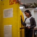 A civilian woman in business casual attire holds a clipboard and inspects or reviews a large yellow flammable materials storage cabinet. A person in military camouflage uniform (OCP/MultiCam pattern) is partially visible to the right, appearing to assist by opening or pointing to the cabinet. The setting appears to be a military facility or warehouse undergoing an inspection or inventory check.