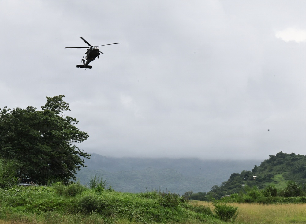 A military helicopter, consistent with a UH-60 Black Hawk, is flying low over a lush tropical or subtropical landscape under overcast skies. The terrain features dense green vegetation, rolling hills partially obscured by low clouds or mist, and an open grassy field in the foreground. The scene suggests a military operation, training exercise, or humanitarian mission in a tropical region.