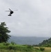 A military helicopter, consistent with a UH-60 Black Hawk, is flying low over a lush tropical or subtropical landscape under overcast skies. The terrain features dense green vegetation, rolling hills partially obscured by low clouds or mist, and an open grassy field in the foreground. The scene suggests a military operation, training exercise, or humanitarian mission in a tropical region.