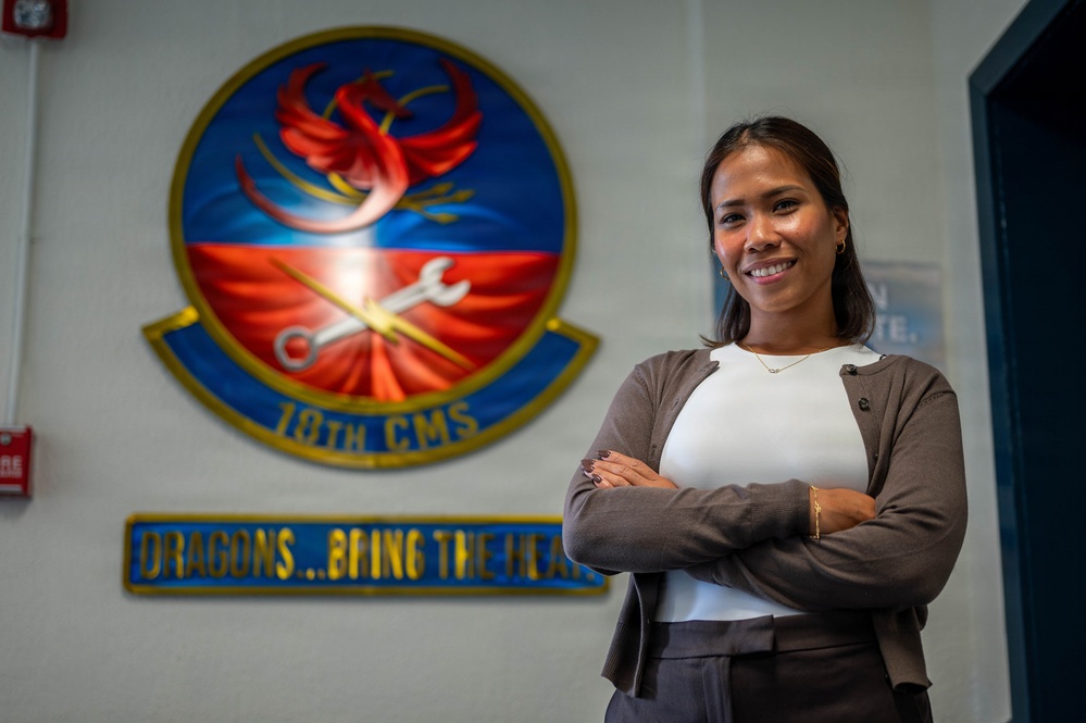 A woman stands smiling with arms crossed in front of a military unit emblem on a wall. The emblem belongs to the 18th CMS (Component Maintenance Squadron), featuring a phoenix with crossed wrench and key symbols. A motto partially visible reads 'DRAGONS...BRING THE HEA[T]'.
