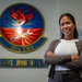 A woman stands smiling with arms crossed in front of a military unit emblem on a wall. The emblem belongs to the 18th CMS (Component Maintenance Squadron), featuring a phoenix with crossed wrench and key symbols. A motto partially visible reads 'DRAGONS...BRING THE HEA[T]'.