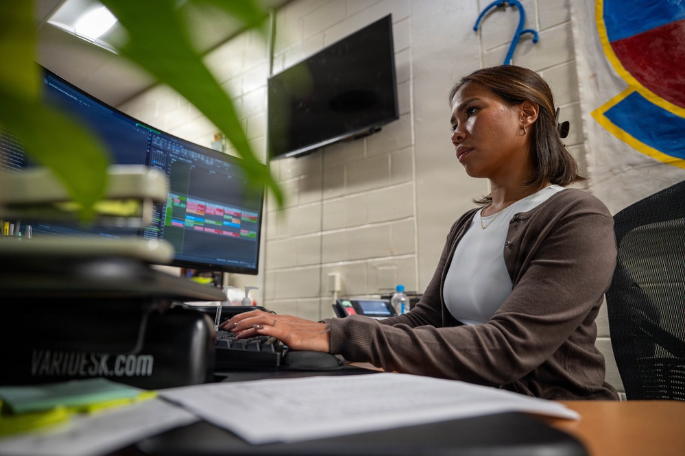 A woman is seated at a desk working on a curved ultrawide monitor displaying what appears to be a scheduling or data management application with colorful blocks. The setting appears to be an office or administrative workspace, likely in a military or institutional environment based on the insignia/emblem visible on the wall. The image is taken from a low angle with a plant in the foreground.