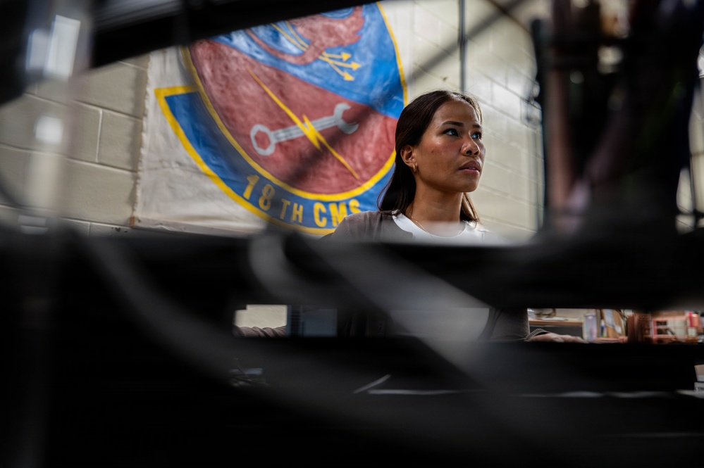 A woman is photographed in what appears to be a military maintenance facility, with the 18th Component Maintenance Squadron (18th CMS) unit banner visible on the wall behind her. The image is taken from a low angle through foreground equipment, creating a dramatic compositional framing. The setting suggests a U.S. Air Force maintenance shop environment.