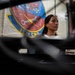 A woman is photographed in what appears to be a military maintenance facility, with the 18th Component Maintenance Squadron (18th CMS) unit banner visible on the wall behind her. The image is taken from a low angle through foreground equipment, creating a dramatic compositional framing. The setting suggests a U.S. Air Force maintenance shop environment.