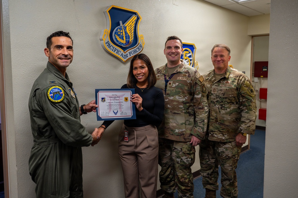 Four individuals pose for a photo in front of a Pacific Air Forces emblem/shield on a wall. A civilian woman is being presented with what appears to be a '15th Wing Shopper of the Week' or similar recognition certificate. Three of the individuals are in military uniform while one is in civilian attire.