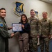 Four individuals pose for a photo in front of a Pacific Air Forces emblem/shield on a wall. A civilian woman is being presented with what appears to be a '15th Wing Shopper of the Week' or similar recognition certificate. Three of the individuals are in military uniform while one is in civilian attire.
