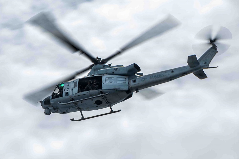 A U.S. Marines military helicopter is captured in flight against an overcast sky. The aircraft displays the number '67' on its nose and 'MARINES' lettering along its fuselage. The main rotor blades and tail rotor are visibly spinning, captured with motion blur indicating active flight.