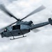 A U.S. Marines military helicopter is captured in flight against an overcast sky. The aircraft displays the number '67' on its nose and 'MARINES' lettering along its fuselage. The main rotor blades and tail rotor are visibly spinning, captured with motion blur indicating active flight.