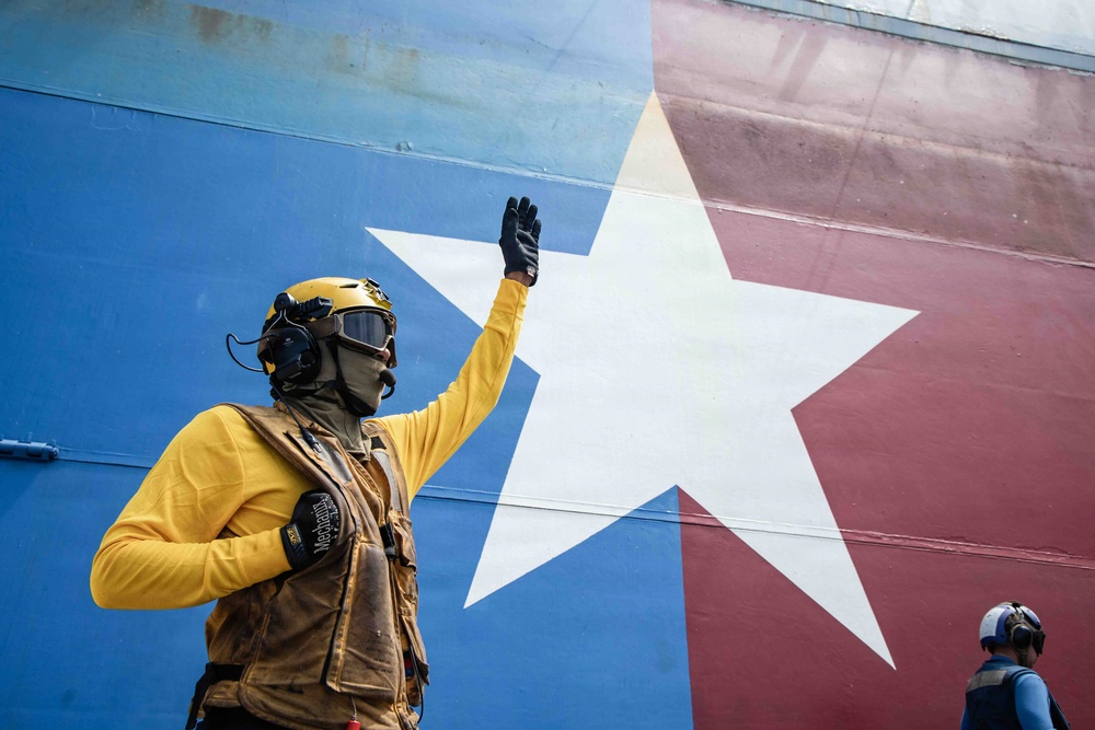 A military or naval deck crew member wearing a yellow jersey, tactical vest, helmet with goggles, and black gloves raises one hand in a signaling gesture. Behind them is a large painted surface featuring a white star on a blue and red background, consistent with the Texas state flag design. A second person wearing a blue helmet and ear protection is partially visible in the lower right corner.
