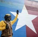 A military or naval deck crew member wearing a yellow jersey, tactical vest, helmet with goggles, and black gloves raises one hand in a signaling gesture. Behind them is a large painted surface featuring a white star on a blue and red background, consistent with the Texas state flag design. A second person wearing a blue helmet and ear protection is partially visible in the lower right corner.