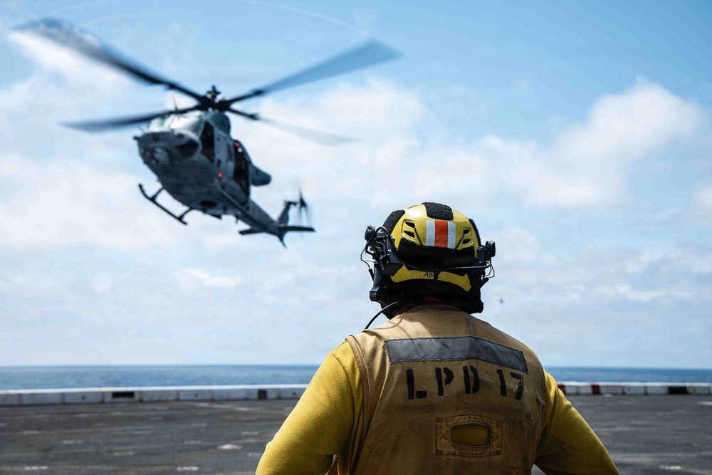 A naval flight deck crew member wearing a yellow jersey and helmet marked 'LPD 17' observes a military helicopter during flight operations on a ship's deck. The helicopter, appearing to be a UH-1Y Venom or similar twin-rotor utility helicopter in gray military livery, is either landing or departing the flight deck. The scene takes place at sea under partly cloudy skies.