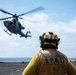 A naval flight deck crew member wearing a yellow jersey and helmet marked 'LPD 17' observes a military helicopter during flight operations on a ship's deck. The helicopter, appearing to be a UH-1Y Venom or similar twin-rotor utility helicopter in gray military livery, is either landing or departing the flight deck. The scene takes place at sea under partly cloudy skies.