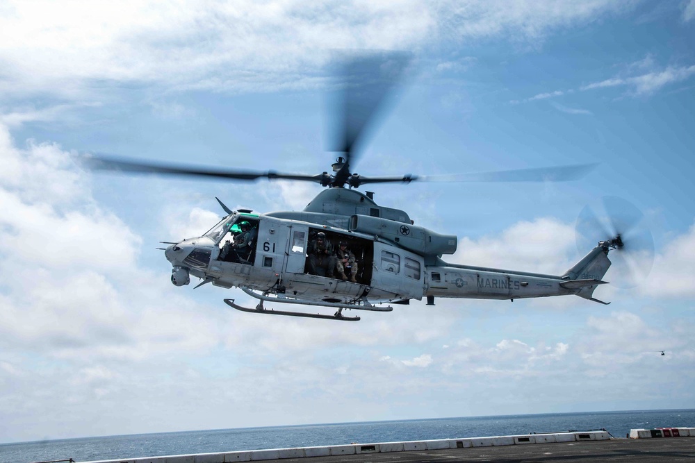 A U.S. Marines UH-1Y Venom helicopter (tail number 61) is captured in flight, lifting off or hovering over what appears to be the deck of a naval vessel at sea. The helicopter's side door is open with crew members visible inside, and the main rotor blades show motion blur indicating active flight. A second helicopter is faintly visible in the distant background.
