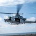 A U.S. Marines UH-1Y Venom helicopter (tail number 61) is captured in flight, lifting off or hovering over what appears to be the deck of a naval vessel at sea. The helicopter's side door is open with crew members visible inside, and the main rotor blades show motion blur indicating active flight. A second helicopter is faintly visible in the distant background.