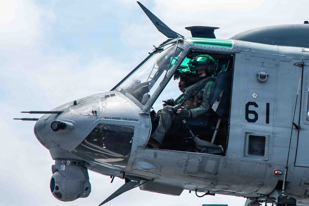 A close-up photograph of a military attack helicopter, specifically appearing to be a Bell AH-1Z Viper, with bureau number 61 visible on the fuselage. Two crew members in flight suits and helmets with night-vision equipment are visible in the tandem cockpit. The aircraft is airborne and the name 'CAPT J.J. BALLERINO TOLENTINO' is stenciled on the cockpit frame.