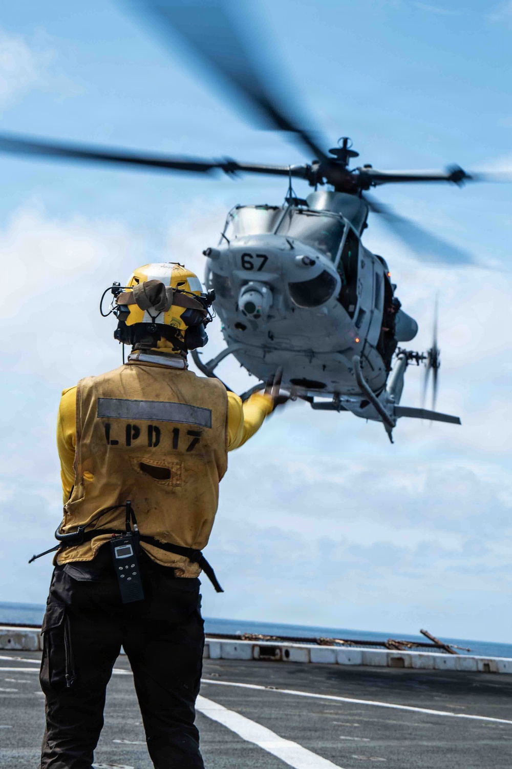 A naval flight deck crew member wearing a yellow jersey marked 'LPD17' signals a military helicopter numbered '67' during flight deck operations aboard a U.S. Navy amphibious transport dock ship. The crew member is equipped with a helmet, hearing protection, and a radio, and is directing the helicopter with an outstretched arm. The scene takes place on an open flight deck over open ocean.