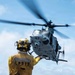 A naval flight deck crew member wearing a yellow jersey marked 'LPD17' signals a military helicopter numbered '67' during flight deck operations aboard a U.S. Navy amphibious transport dock ship. The crew member is equipped with a helmet, hearing protection, and a radio, and is directing the helicopter with an outstretched arm. The scene takes place on an open flight deck over open ocean.