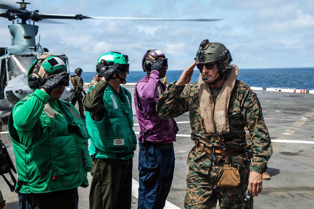 Military personnel are gathered on a naval vessel's flight deck, with several individuals in colored safety vests and helmets rendering salutes to a figure in USMC digital camouflage uniform. A helicopter is visible in the background on the left side of the deck. The scene appears to depict a formal greeting or arrival ceremony at sea.