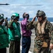 Military personnel are gathered on a naval vessel's flight deck, with several individuals in colored safety vests and helmets rendering salutes to a figure in USMC digital camouflage uniform. A helicopter is visible in the background on the left side of the deck. The scene appears to depict a formal greeting or arrival ceremony at sea.